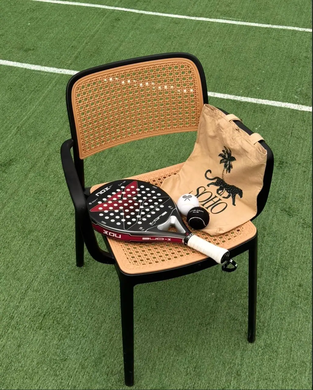 Padel racket and Soho Boulevard tote bag resting on a rattan chair beside the court