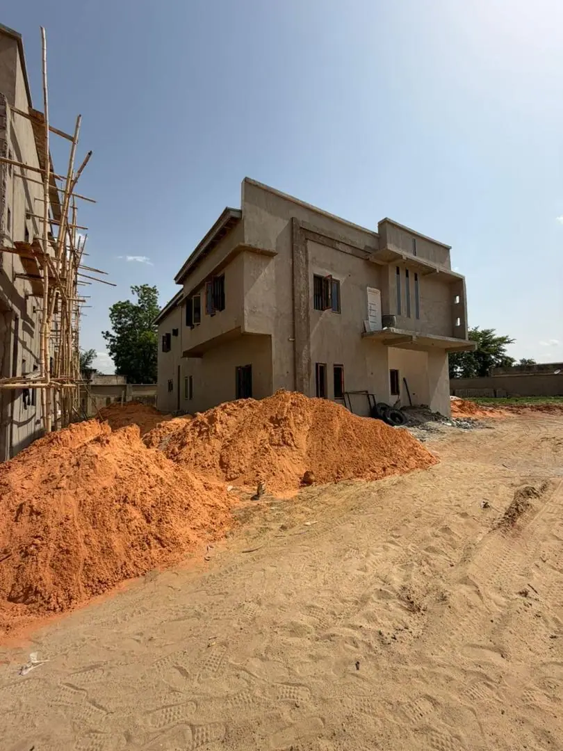 Site photograph of a two-storey Three of Us duplex shell with timber scaffolding and mounds of laterite soil